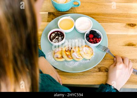 Giovane donna che fa colazione - pancake al formaggio caserma e caffè cappuccino su tavolo di legno in caffetteria. Vista dall'alto Foto Stock