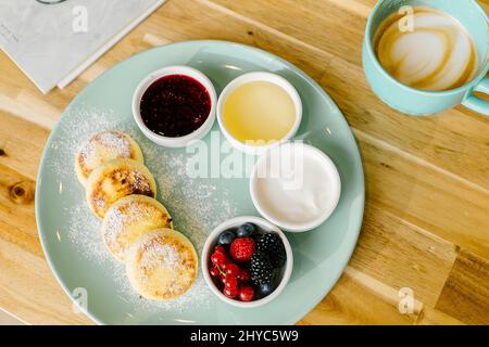 Pancake al formaggio casolare o scirniki per colazione con frutti di bosco freschi e cappuccino caffè su tavola di legno. Vista dall'alto Foto Stock