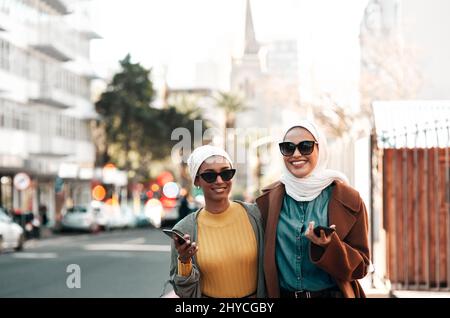 Questa è la generazione millenaria. Scatto corto di due giovani donne musulmane attraenti che indossano occhiali da sole e tengono i loro cellulari mentre la città vista Foto Stock