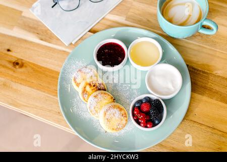 Sana colazione estiva. Pancake al formaggio casolare con frutti di bosco freschi e cappuccino su tavola di legno. Vista dall'alto Foto Stock