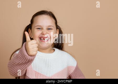 Ritratto di giovane bella ragazza sorridente gioiosa con lunghi capelli scuri in bianco, maglione rosa in piedi, mostrando pollici segno, gesto di approvazione su Foto Stock