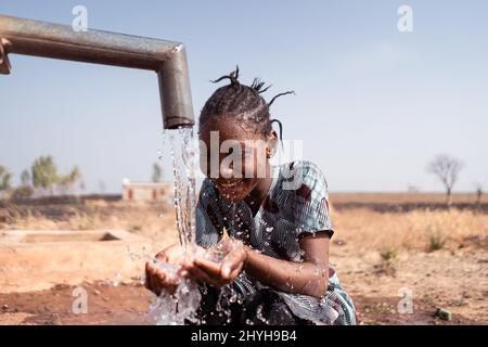 Piccola ragazza africana felice accovacciata davanti ad un pozzo in un ambiente arido da qualche parte nella regione del subsaharn, bevendo lotti di acqua dolce Foto Stock