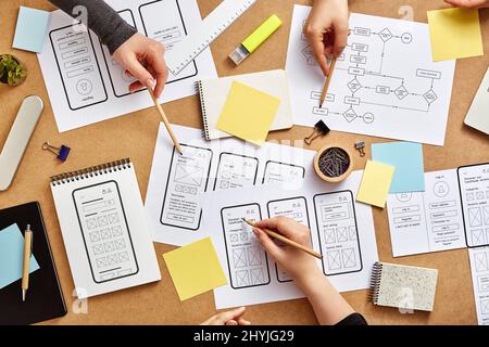 Tre progettisti di Web UX che lavorano insieme a una scrivania occupata con numerosi schizzi di strutture a reticolo per app mobili e flusso di utenti. Vista dall'alto. Foto Stock