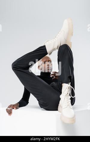 giovane uomo afroamericano in abito elegante posato su cubo bianco isolato su grigio Foto Stock