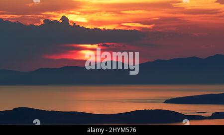 Tramonto, sole grande dietro le nuvole grigie e sopra una catena montuosa, promontori come silhouette in primo piano, mare calmo, cielo arancione-rosso, Palaiopolis, Andros Foto Stock