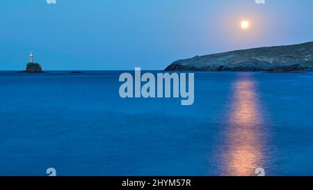 Luna piena, luna sopra costa rocciosa, raggio di luna sul mare, mare calmo, cielo blu e nuvoloso, faro su piccola isola rocciosa brilla, Andros Island Foto Stock