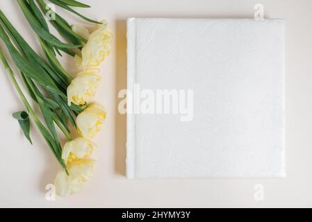 Album fotografico bianco per famiglie o matrimoni con copertina in pelle con spazio vuoto per il testo circondato da tulipani gialli su sfondo chiaro. Foto di famiglia archi Foto Stock