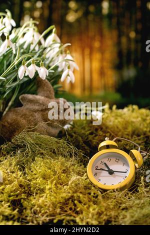 Saluto di Pasqua. Orologio allarme giallo e coniglio coniglietto su muschio nella foresta di primavera, bella fiore bianco nevicate fiori nella foresta selvaggia sullo sfondo. Foto Stock