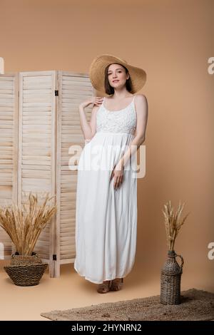 Donna incinta elegante in cappello di paglia in posa sul beige Foto Stock
