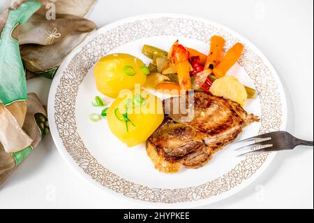 Bistecca di maiale arrosto, patate bollite e verdure cotte su piatto bianco con forchetta, su sfondo bianco con asciugamano, primo piano. Foto Stock