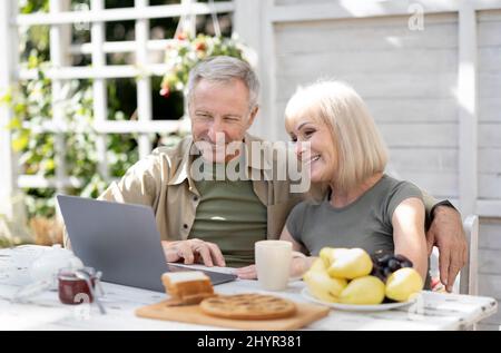 Coppia di anziani felici che usano il computer portatile all'aperto, seduti sulla terrazza di casa e navigare in internet o fare una videochiamata Foto Stock