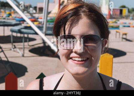 Giovane donna con capelli arancioni neri, occhiali da sole, sorridente, guardando la macchina fotografica mentre si trova di fronte a un parco trampolino a maiorca, da vicino Foto Stock