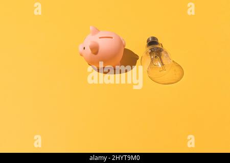 Piggy Bank e lampadina su sfondo giallo. Concetto di elettricità, aumento dei prezzi, impoverimento ed economia. Foto Stock