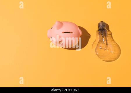 Piggy Bank e lampadina su sfondo giallo. Concetto di elettricità, aumento dei prezzi, impoverimento ed economia. Foto Stock