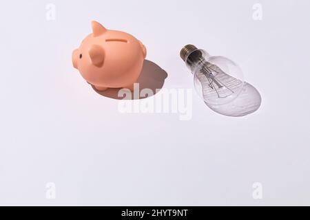 Piggy banca e lampadina su sfondo bianco concetto di elettricità, aumento dei prezzi, impoverimento ed economia. Foto Stock