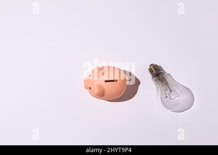 Piggy banca e lampadina su sfondo bianco concetto di elettricità, aumento dei prezzi, impoverimento ed economia. Foto Stock