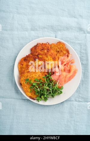 Frittelle di patate. Frittelle di patate fatte in casa fritte o latkes con crema, cipolle verdi, microgreens, salmone rosso e salsa in piatto rustico su lino blu ta Foto Stock