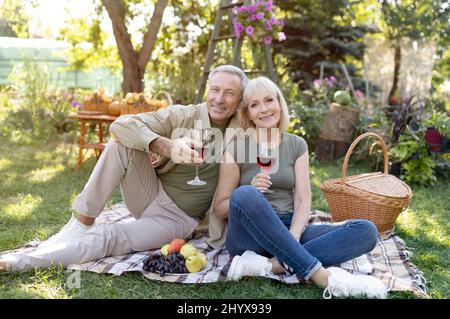 Pensionamento spensierato. Sposi anziani felici che bevono vino, seduti su coperta mentre si fanno pic-nic nel loro giardino Foto Stock