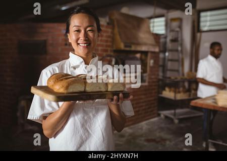 Ritratto di sorridente panettiere asiatico di metà adulta femmina che tiene tavola da servire con pane fresco Foto Stock