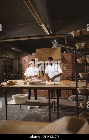 Panettiere multirazziale maschile e femminile per adulti di media età che parlano mentre impastano la pasta al tavolo in panetteria Foto Stock