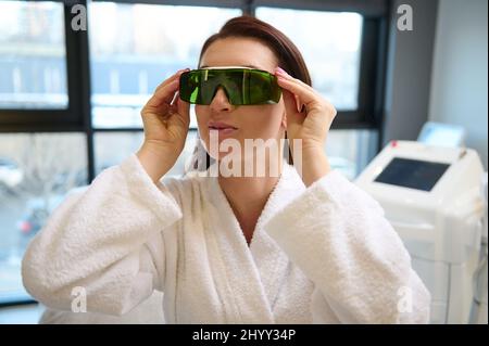 Donna europea sicura di mezza età in accappatoio bianco mette su occhiali protettivi UV, pronto per il trattamento di bellezza con apparecchi laser in un benessere Foto Stock