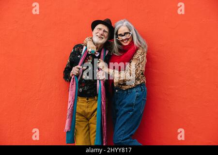 Spensierate la coppia anziana ridendo mentre si alza insieme su uno sfondo rosso. Coppia allegra e anziana che indossa abiti colorati casual. Buon tappetino Foto Stock