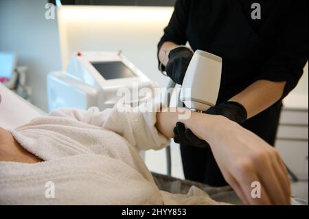 Primo piano di un estetista cosmetologo irriconoscibile che esegue una procedura di rimozione dei capelli da parte di una donna con moderne apparecchiature laser in wel Foto Stock