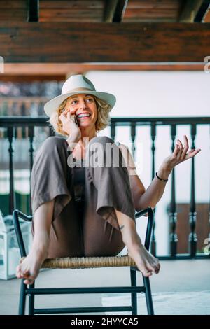 Ritratto di una giovane bionda caucasica matura nel suo 50s parlando su un telefono cellulare sorridendo e indossando un cappello e seduto in una posa rilassata in un ro Foto Stock
