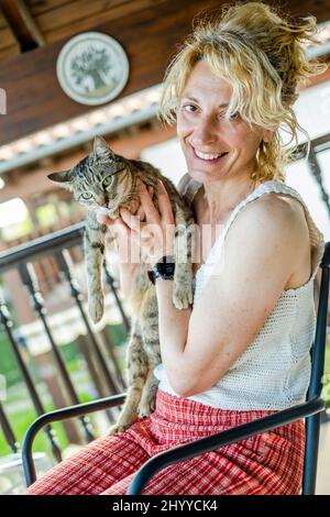 ritratto di una giovane bionda caucasica matura che sorride con il gatto a casa sul portico seduto su una sedia a dondolo. Concetto di stile di vita. Foto Stock