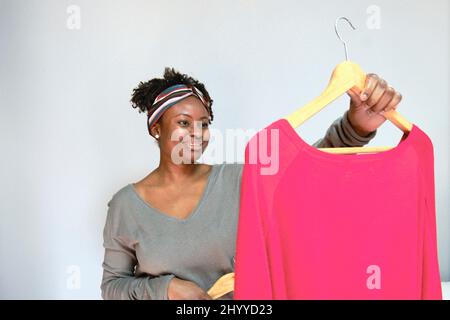 Ritratto di giovane donna nera con acconciatura afro con abiti su appendiabiti interni a casa. Concetto di stile di vita. Foto Stock