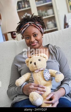 Ritratto di una giovane donna nera con acconciatura afro e un orsacchiotto al coperto seduto su un divano. Concetto di stile di vita. Foto Stock