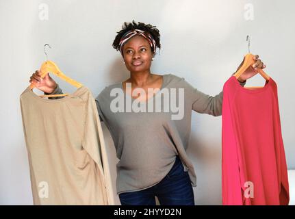 Ritratto di giovane donna nera con acconciatura afro con abiti su appendiabiti interni a casa. Concetto di stile di vita. Foto Stock