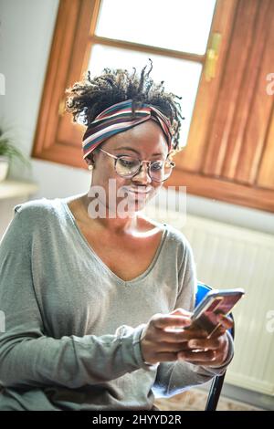 Giovane donna nera con acconciatura afro e occhiali con un telefono cellulare interno in una casa. Concetto di stile di vita. Foto Stock