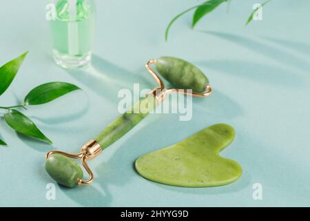 Rullo massaggiante per il viso con due teste e raschietto gua sha di pietra di giada verde su sfondo azzurro. Massaggio viso di bellezza, SPA self c Foto Stock