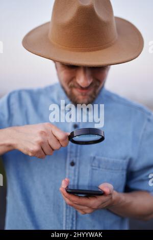 Uomo indossando jeans camicia e cappello guardando con loupe sullo smartphone come detective, investigatore o concetto di ricerca. Uomo caucasico bearded che guarda con lente d'ingrandimento allo smartphone. Immagine di alta qualità Foto Stock