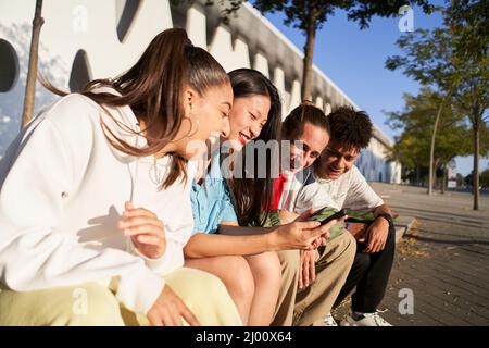 Gruppo di giovani alla moda che chiacchierano insieme seduti all'aperto. Gli studenti si divertono insieme. Ragazze e ragazzi che usano il telefono e sorridono Foto Stock