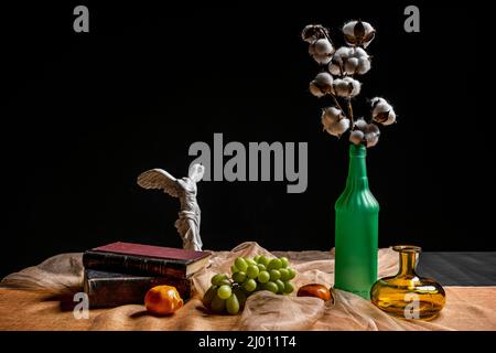 Ancora vita con un ramo di cotone in una bottiglia di vetro con uva bianca, vasetti di vetro e libri antichi Foto Stock