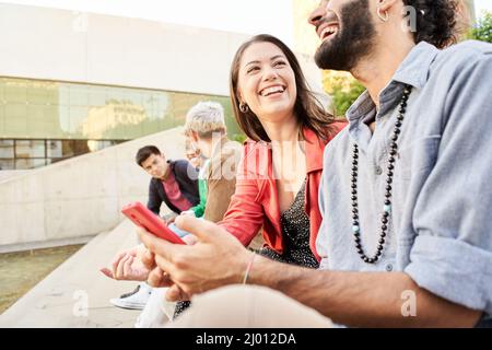 Giovane coppia che usa smartphone digitali nel campus universitario. Gruppo di amici che guardano i cellulari condividere contenuti e ridere. Giovani, amicizia e. Foto Stock