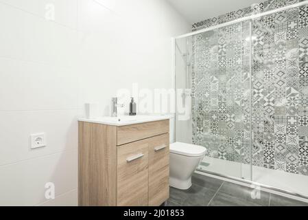 bagno con sanitari in porcellana bianca, cabina doccia con parete piastrellata con piastrelle idrauliche e pareti bianche, mobili in legno e pavimento in gres scuro Foto Stock