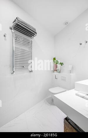 bagno con finiture in porcellana bianca, pareti in piastrelle bianche e pavimenti in marmo e radiatore asciugamani in acciaio inox Foto Stock