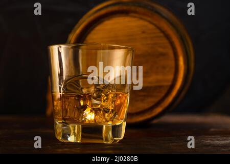 Un bicchiere di whiskey e ghiaccio si trova sulle tavole di quercia sullo sfondo di un barile di quercia. Primo piano. Whiskey da una distilleria domestica Foto Stock