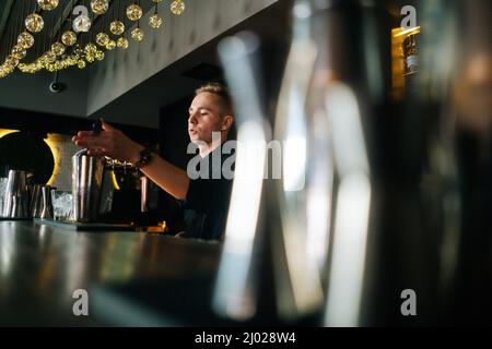 Vista laterale ad angolo basso del barman fiducioso che fa un rinfrescante cocktail alcolico in piedi dietro il bancone del bar nel moderno e scuro nightclub, Foto Stock