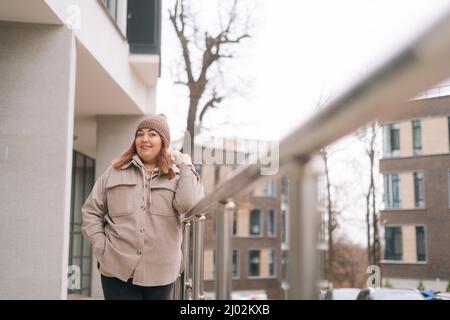Ritratto di donna felice sovrappeso in caldo cappello e giacca in piedi vicino ringhiera di ufficio edificio in strada della città in nuvoloso giorno d'autunno Foto Stock