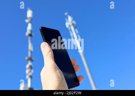 Smartphone in mano sullo sfondo delle torri cellulari. Apparecchiature per telecomunicazioni mobili, potenza del segnale telefonico Foto Stock