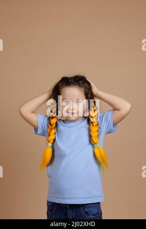 Offeso piccola ragazza che ha sbagliato testa di tenuta con le mani guardando fotocamera con aspetto sullen che hanno kanekalon trecce di colore giallo in blu t-shirt on Foto Stock