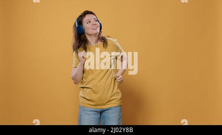 Ritratto di donna casual che ascolta musica allegra con cuffie wireless e che fa mosse di danza stravaganti in studio. Donna positiva piena di energia con auricolari che hanno partito e ballare da soli. Foto Stock