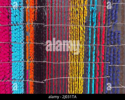 Narayanganj, Dhaka, Bangladesh. 16th Mar 2022. I lavoratori appendono migliaia di diversi tessuti colorati su fili di ferro legati tra un telaio di bambù e costantemente li trasformano in modo che si asciughino perfettamente in campo allagato a Narayanganj, Bangladesh. I fili di ferro sono usati fra una struttura di bambù per generare le linee giganti di lavaggio per la parte finale del processo di tintura mentre i tessuti sono asciugati nel sole. I fili luminosi di panni blu, rosa, arancione e verde-tinti appendono sopra il campo erboso in una rete abbagliante di colori interbloccanti. Questa è la parte finale del processo di tintura dopo la quale il panno è Foto Stock