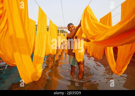 Narayanganj, Dhaka, Bangladesh. 16th Mar 2022. I lavoratori appendono migliaia di diversi tessuti colorati su fili di ferro legati tra un telaio di bambù e costantemente li trasformano in modo che si asciughino perfettamente in campo allagato a Narayanganj, Bangladesh. I fili di ferro sono usati fra una struttura di bambù per generare le linee giganti di lavaggio per la parte finale del processo di tintura mentre i tessuti sono asciugati nel sole. I fili luminosi di panni blu, rosa, arancione e verde-tinti appendono sopra il campo erboso in una rete abbagliante di colori interbloccanti. Questa è la parte finale del processo di tintura dopo la quale il panno è Foto Stock