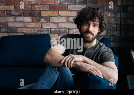 Ritratto di ragazzo fiducioso bearded sorridente guardando la macchina fotografica mentre si siede su un comodo divano in salotto accogliente con parete di mattoni. Foto Stock