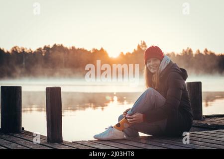 Giovane brunetta con un bel sorriso realistico siede su un ponte di legno accanto ad un lago all'alba. Cappello rosso con cappotto nero e scarpe bianche. Stile di moda. Foto Stock